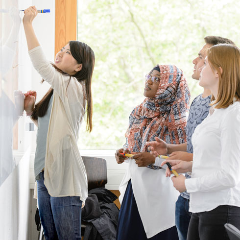 Gruppe von Studierenden (unterschiedlicher Nationalität) vor einem Whiteboard, eine asiatische Studentin schreibt etwas an, die anderen schauen interessiert zu.