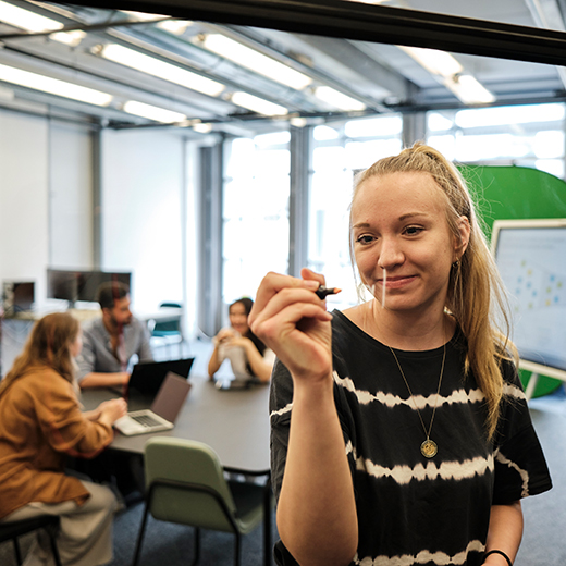 Studentin nutzt transparente Tafel, im Hintergrund arbeitende Projektgruppe