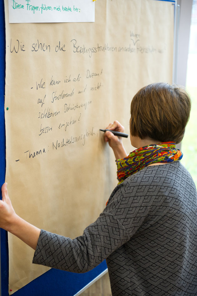 Eine Frau (von hinten fotografiert) schreibt auf ein großes Blatt Papier, das an einer Wand hängt.