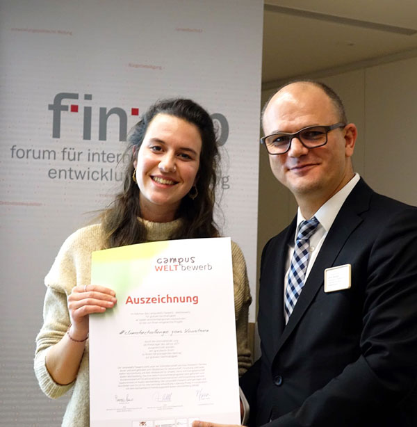 Eine Frau und ein Mann posieren mit einer Urkunde in den Händen fürs Foto. Lesbar sind die Worte "Auszeichnung" und das Logo "campus WELTbewerb".