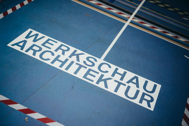 Die Aufschrift "Werkschau Architektur" klebt auf einem Boden. Rundherum klebt rot-weißes Absperrband.