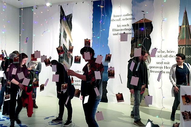 (W)ortverliebt in Konstanz: Ausstellung zu von Wolkenstein Menschen in einem Ausstellungsraum: Von der Decke hängen Fotos, an der Wand dahinter kleben abgecshnittene Bildausschnitte von Gebäuden. Dazwischen stehen Texte.
