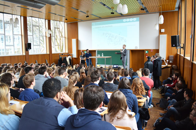 Ein überbesetzter Vorlesungsraum der HTWG. Drei Sprecher*innen halten eine Präsentation.