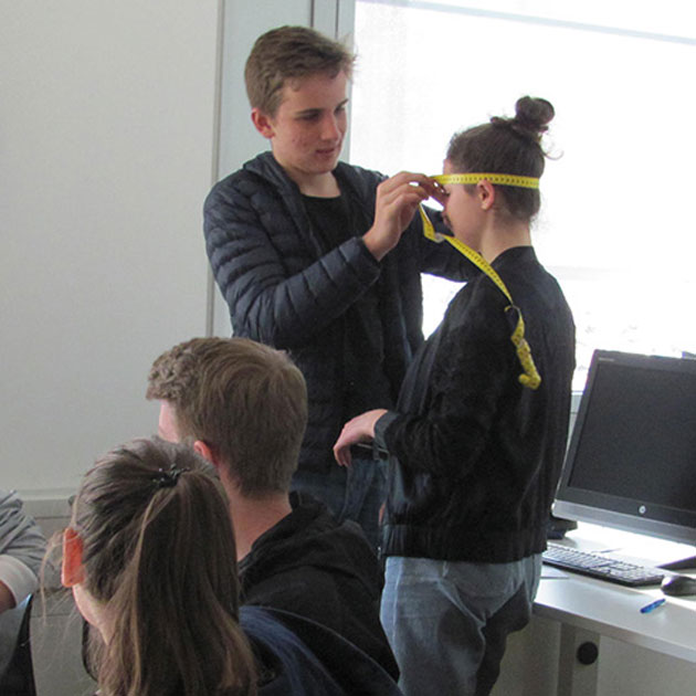 Ein junger Mann in einem Seminarraum misst den Kopfumfang einer jungen Frau mit einem Maßband.