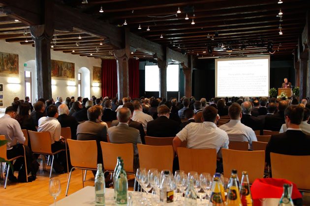 Ein großer Präsentationssaal voller Menschen von hinten fotografiert. Vorne spricht ein Mann vor einer Leinwand hinter einem Rednerpult. Im Bildvordergrund steht ein Tisch mit Flaschen und Gläsern.