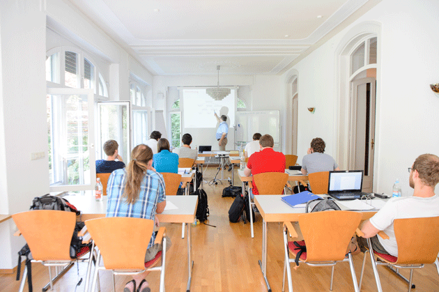 Ein Seminarraum voller Studierender von hinten fotografiert. Vorne zeigt ein Mann auf etwas auf einer Leinwand. 