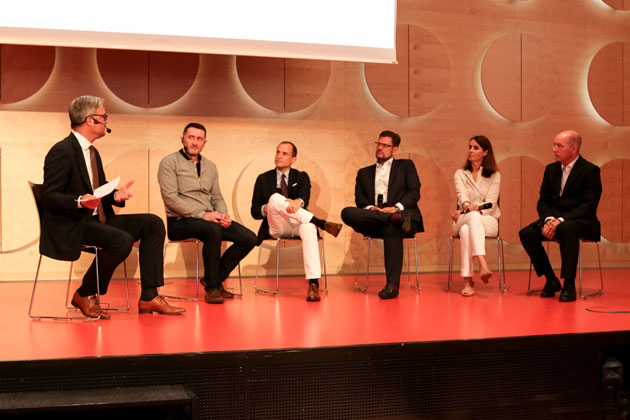 Ein Moderator und fünf weitere Personen sitzen auf einer Bühne.