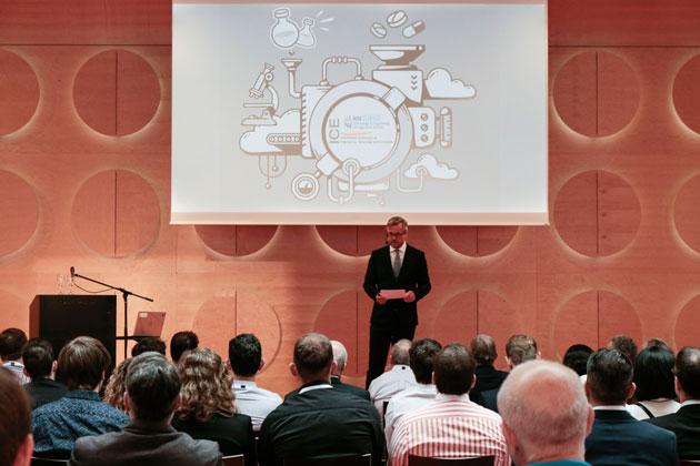 Ein Moderator steht vor Publikum auf einer Bühne. Hinter ihm hängt eine Leinwand. Darauf abgebildet ist die Illustration einer Maschine.