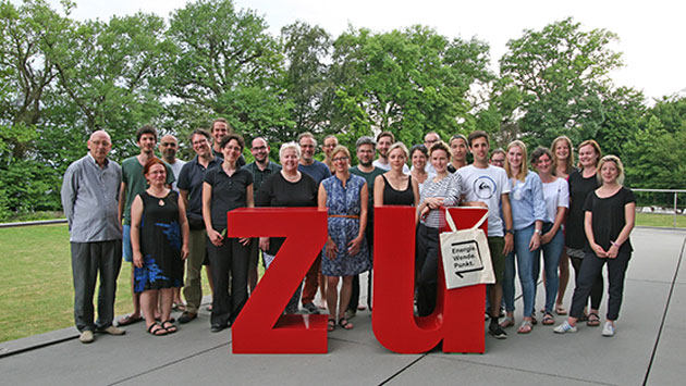 Eine große Gruppe Menschen posiert im Freien hinter zwei großen Buchstaben ("Z" und "U") fürs Foto.
