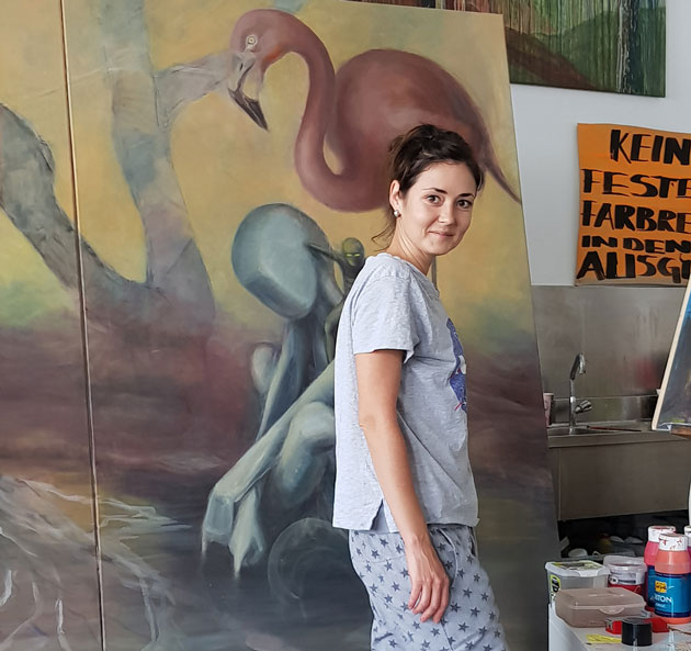 Eine junge Frau steht in einem Atelier vor einem großen Bild in blassen Farben. Es zeigt ein Flamingo und zwei menschliche Figuren in unterschiedlichen Größen.