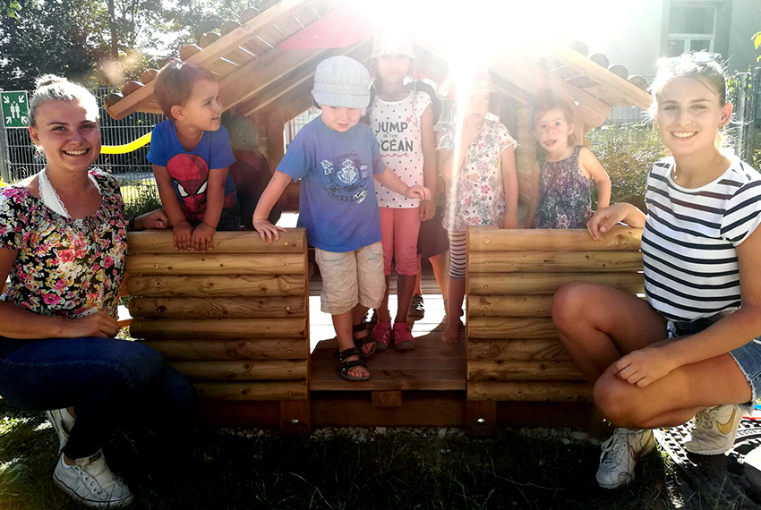 Zwei junge Frauen posieren kniend vor einem kleinen Holzhaus für Kinder fürs Foto. Im Haus stehen fünf Kinder.