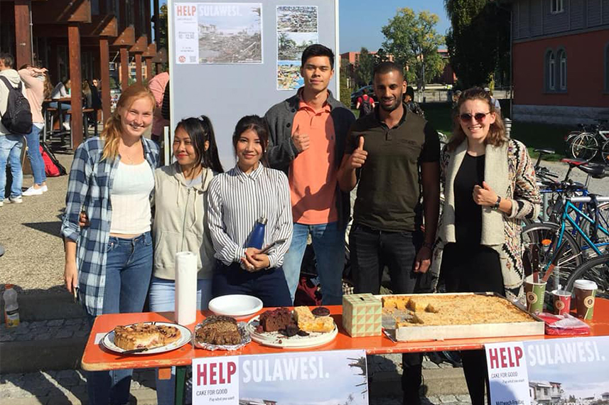 Sechs junge Menschen stehen hinter einem Biertisch voller Kuchen. An der Tischkante und auf einer Infotafel hinter ihnen hängen Plakate mit der Aufschrift "Help Sulawesi".