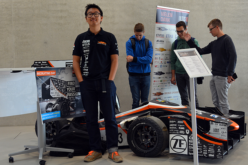 Ein junger Mann in Teamkleidung steht vor dem Rennwagen des Bodensee Racing Teams der HTWG, einer Infotafel und einer Tafel mit Sponsoren. Im Hintergrund betrachten drei junge Männer den Rennwagen und sprechen miteinander.