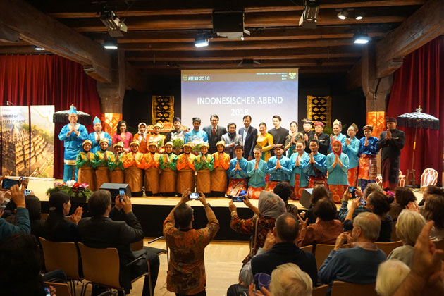Eine große Gruppe Menschen in traditioneller indonesischer Kleidung auf einer Bühne vor Publikum. Auf einer Leinwand hinter ihnen steht "Indonesischer Abend 2018".