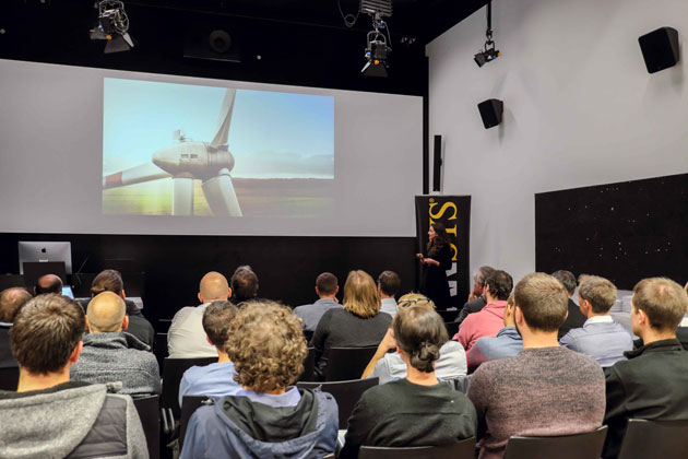 In einem Veranstaltungssaal sitzt das Publikum in Kinobestuhlung und blickt auf eine Leinwand, auf der die Turbine eines Windrads zu sehen ist.