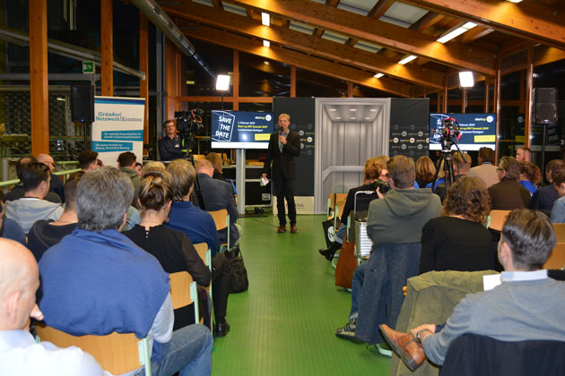 In einem Raum mit grünem Boden und Holzbalken sitzen rund 50 Personen mit Blickrichtung auf Prof. Dr. Oliver Haase, der vor einem Bildschirm in ein Mikrofon spricht. 