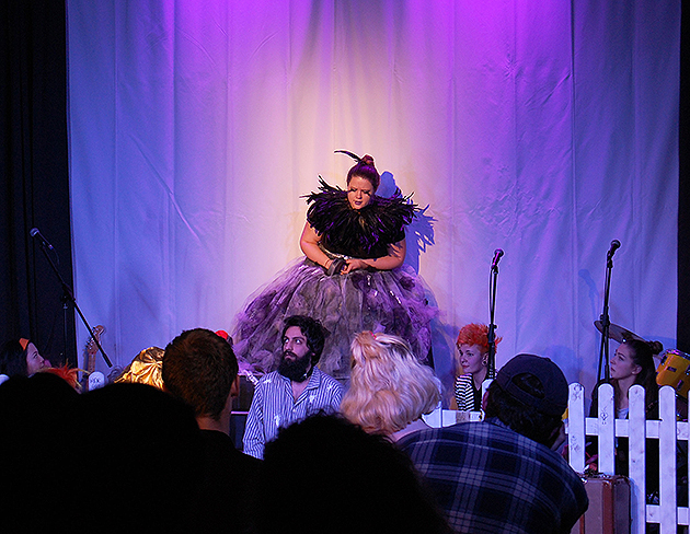Auf einer lila ausgeläuchteten Bühne steht ein Frau in einem schwarzen Federkleid.