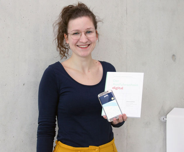 Vanessa Schätzle steht vor einer Betonwand. Mit der linken Hand hält sie DinA-4-Papiere und ein Smartphone vor ihren Oberkörper.