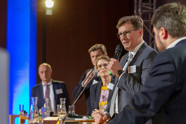 Prof. Dr. Carsten Manz steht auf einem Podium und spricht in ein Mikrofon, das er in der linken Hand hält. Eine Frau rechts von ihm und drei weitere Männer auf dem Podium blicken zu ihm.