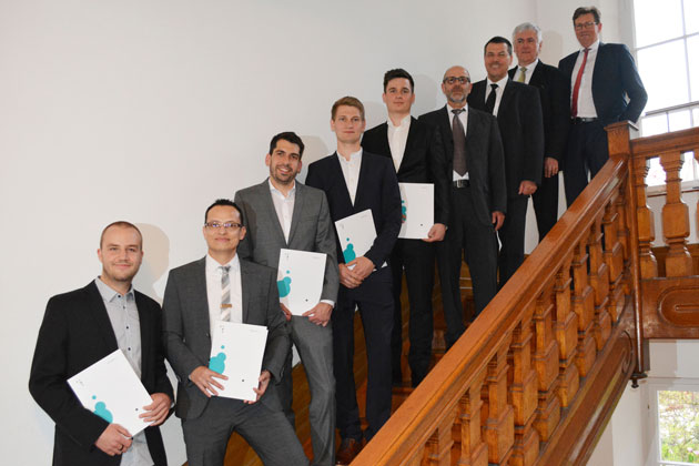 9 Männer in Anzügen stehen auf einer Treppe. Sie halten jeweils eine Mappe mit HTWG-Logo in der Hand und lächeln in Richtung des Fotografen.