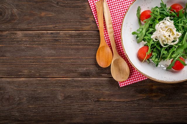 Blick aus der Vogelperspektive auf einen Holztisch, auf dem auf einem rot-weiß-kariertem Tuch ein Teller mit Salat neben zwei Kochlöffeln steht.