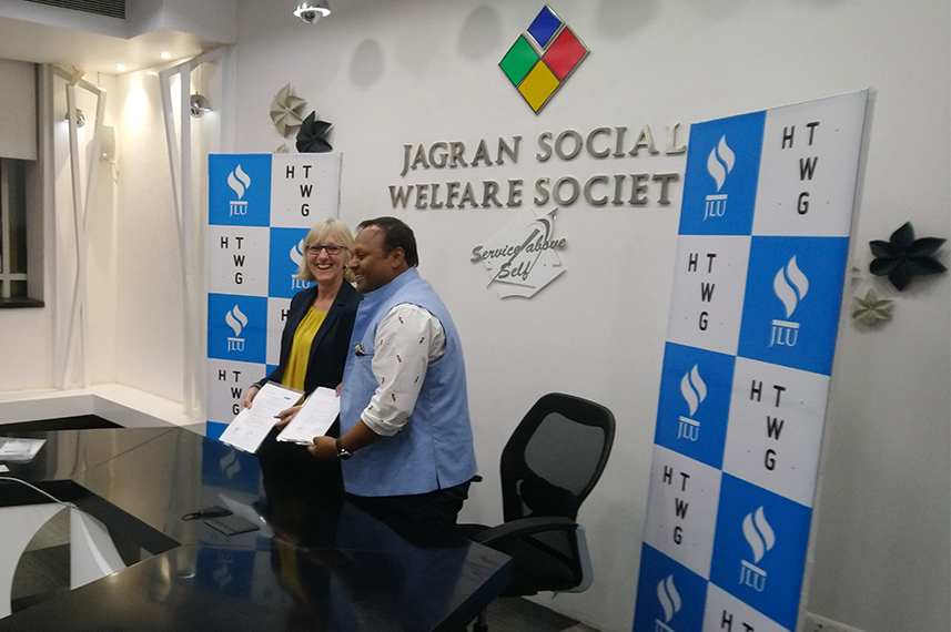 Prof. Dr. Beate Bergé und Prochancellor Abhishek Mohan Gupta (JLU) stehen vor einer Stellwand. Auf der Stellwand steht Jagran Social Welfare Society. Rechts und links der Stellwand stehen Roll-ups mit den Logos der HTWG und der JLU.