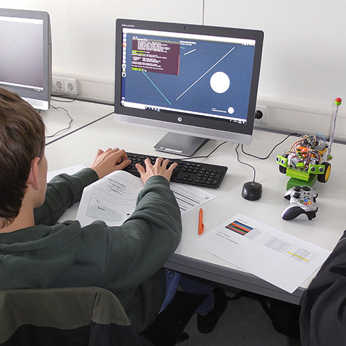 Ein Schüler sitzt an einem Schreibtisch vor einem Bildschirm und schreibt an der Tastatur. Neben ihm liegt ein kleiner Roboter.