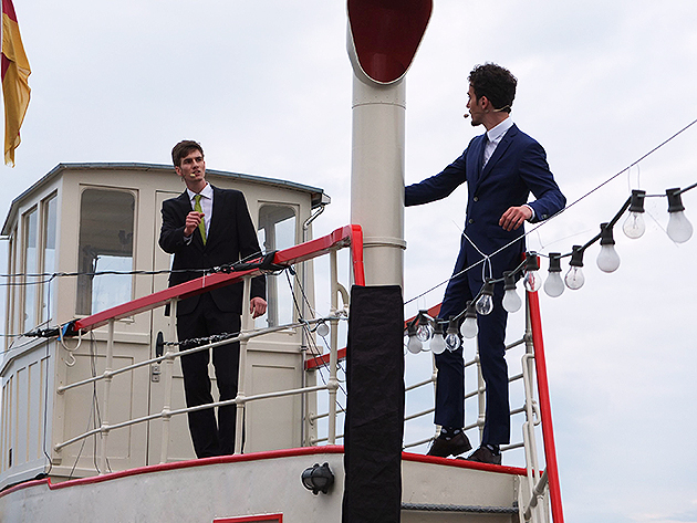 Zwei Studenten in dunklen Anzügen und weißen Hemden stehen sich zugewandt auf der Reling eines Fährschiffs.