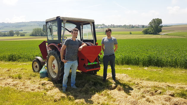 Zwei Studenten stehen vor einem Traktor mit angehängtem Granulatstreuer auf einem grünem Acker.