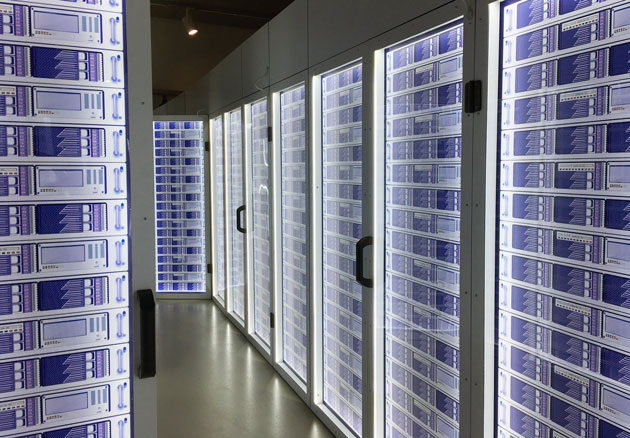 Blick in einen Serverraum. Rechts und links befinden sich hohe Türen, hinter denen sich blau leuchtende Geräte befinden.
