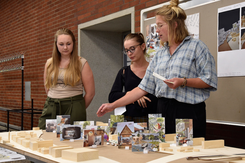Drei Studentinnen stehen hinter einem Tisch, auf dem Architekturentwürfe aufgebaut sind.