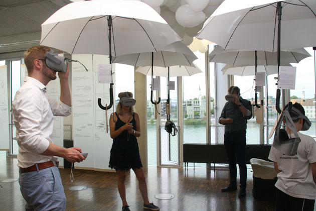 Ein Besucher und zwei Besucherinnen tragen VR-Brillen und stehen neben und unter aufgespannten weißen Regenschirmen in einem Raum.