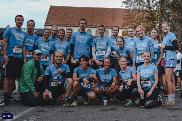 Eine Gruppe von Läuferinnen und Läufern in blauen HTWG-T-Shirts blickt in zwei Reihen aufgestellt in die Kamera. Herbert Rapp kniet in einer grünen Jacke links daneben.