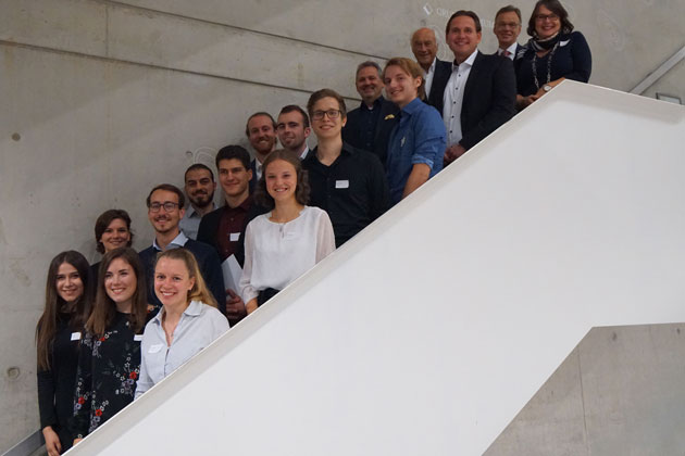 Eine Gruppe von Studentinnen und Studenten sowie Förderer stehen auf einer Treppe. Das Foto zeigt die Gruppe von der Seite mit dem weißen Treppengeländer.