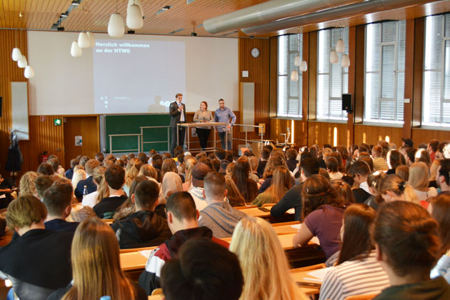 Prof. Dr. Carsten Manz steht mit zwei AStA-Vertretern auf einer Bühne und blickt in das Publikum, von dem die Rücken und Hinterköpfe zu sehen sind.