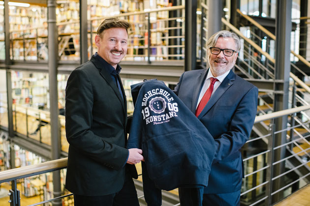 Der Hochschulratsvorsitzende Dr. Stefan Keh steht rechts im Bild und überreicht dem zum Präsidenten gewählten Prof. Dr. Markus Rhomberg einen HTWG-Kapuzenpulli. Im Hintergrund sind Regale der HTWG-Bibliothek zu sehen