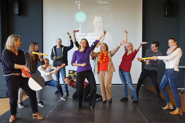 Eine Gruppe von acht HTWG-Mitarbeiterinnnen und -Mitarbeitern bildet mit verschiedenen Körperhaltungen auf einer Bühne ein Standbild zum Thema "Begeisterung auf Knopfdruck"
