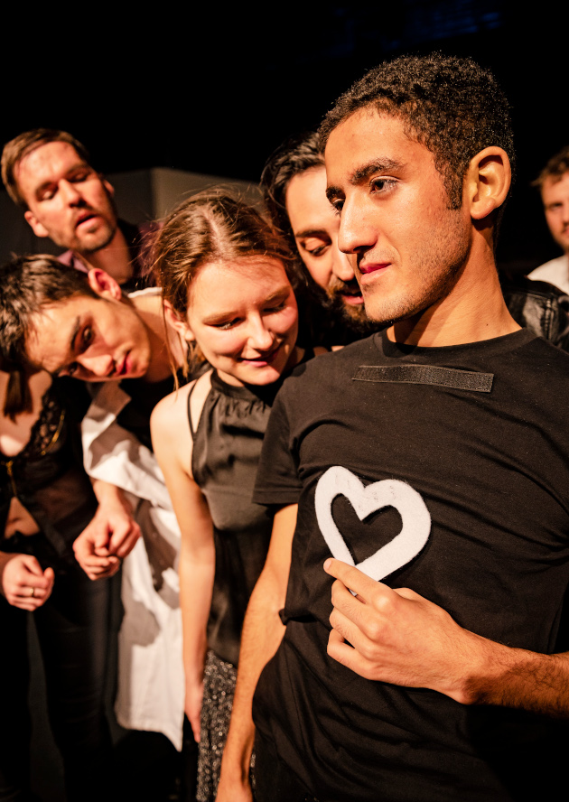 Schauspielerinnen und Schauspieler steht eng nebeneinander. Sie blicken auf das weiße Herz auf dem schwarzen T-Shirt eines der Schauspieler.