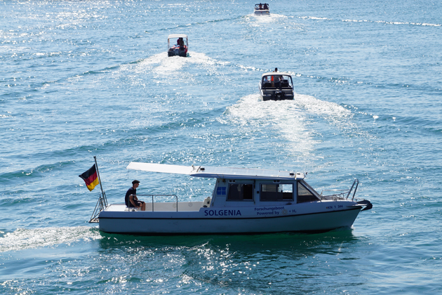 Auf einer Wasserfläche ist im Vordergrund ein weißes Boot von der Seite zu sehen. Hinter dem Boot sind weitere drei Boote zu sehen.