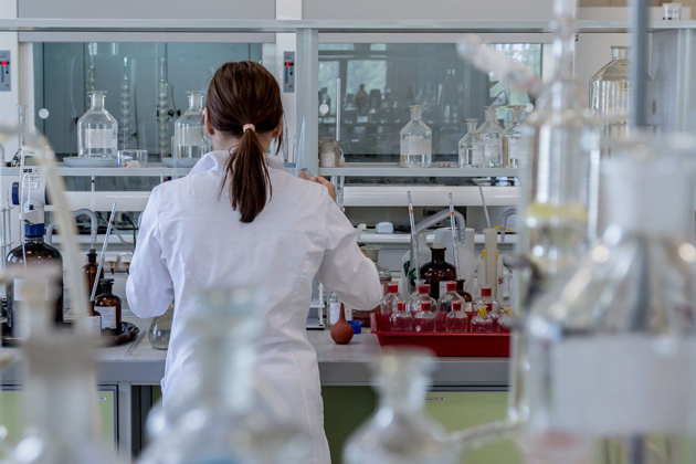Zu sehen ist eine Frau in einem weißen Laborkittel von hinten. Sie steht in einem Chemielabor an einem Arbeitstisch.