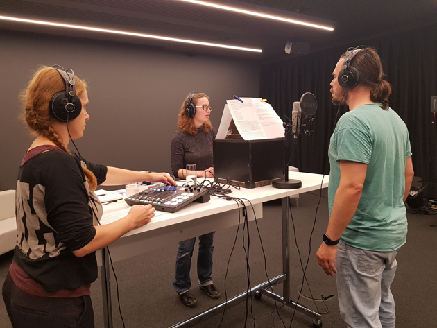 Szene im Tonstudio. Um einen Tisch stehen von links Theaterpädagogin Anna Hertz, Studentin Janina, Schauspieler Thomas Fritz Jung. Sie alle tragen Kopfhörer und blicken auf Textseiten vor sich.