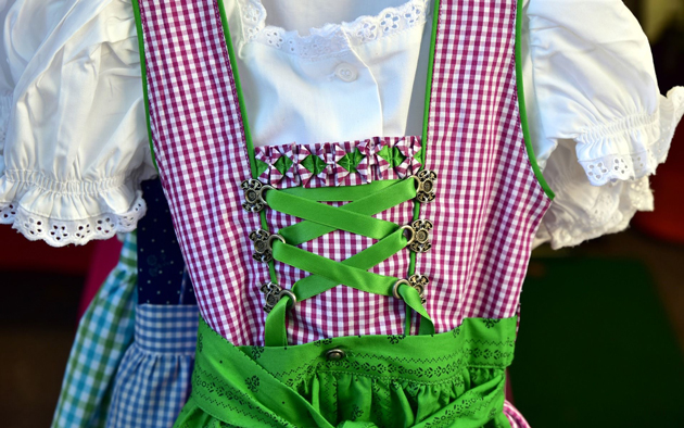 Ein Dirndl hängt an einem Kleiderbügel. Im Bild ist das Oberteil des rot-weiß-karierten Dirndls mit einer grünen Schürze und einer weißen Kurz-Arm-Bluse zu sehen.