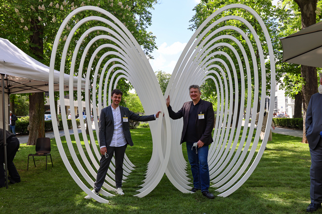 Auf einer grünen Wiese steht ein Denkmal, das mit seinen zwei Spiralen wie die zwei Flügel eines Schmetterlings wirkt. Vor dem linken Flügel steht Michael Wezstein, vor dem rechten Flügel Ulf Meyer.