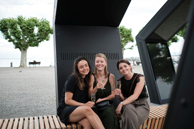 Drei Studentinnen sitzen mit Sektgläsern auf einer Holzbank, die sich durch drei schwarze aufgestellte Fünfecke zieht. Sie halten Sektgläser in den Händen und lachen in die Kamera.