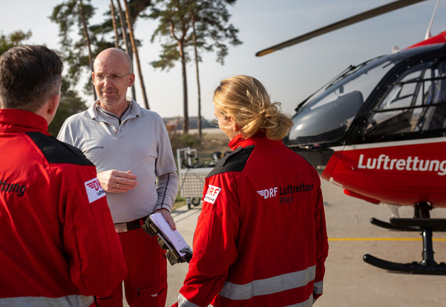 Zwei Männer und eine Frau in roten DRF-Luftrettungsanzügen stehen in ein Gespräch vertieft vor einem roten Hubschrauber der DRF-Luftrettung