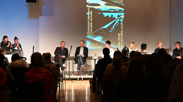 Zwei Männer sitzen mit weiteren Personen auf einem Podium und unterhalten sich. Im Vordergrund sind die Rücken des Publikums zu sehen.