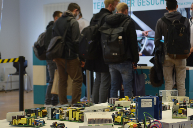 Blick auf einen Messestand, im Hintergrund sind die Rücken von Besuchern zu sehen.