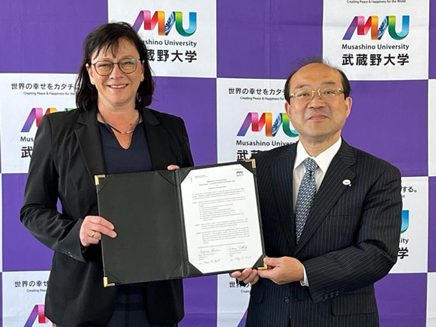 Das Bild zeigt eine Frau und einen Mann, die stolz einen Vertrag in die Kamera halten. Sie stehen vor einer Fotowand der Musashino University.
