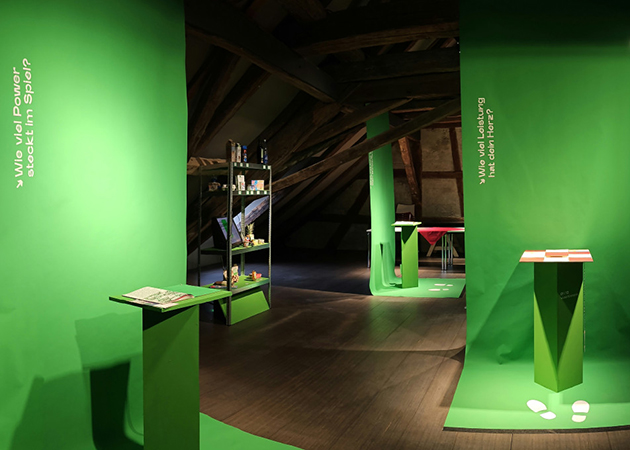 Das Bild zeigt die Ausstellung ohne Menschen. Man sieht grüne Ausstellungswände in einem alten Gebäude mit Holzbalken.