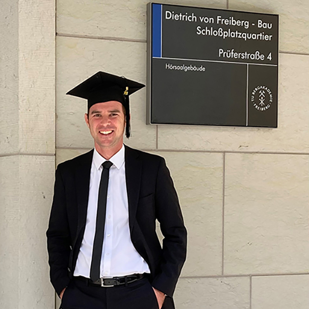 Das Bild zeigt Nico Deistler im schwarzen Anzug und Absolventenhut vor seinem Institutsgebäude.